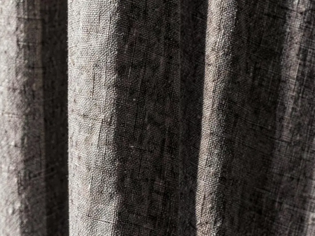 Close médio de tecido de cortina cinza em linho misto, com trama visível e luz lateral criando sombra e profundidade nas dobras